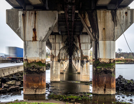 A lowangle view captures the imposing concrete pillars of a highway bridge over calm water. With no people, this atmospheric industrial scene evokes themes of transit, urban decay, and progress.