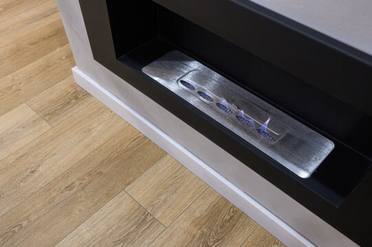 Sleek linear bioethanol burner features brushed-steel insert, multiple oval ports, dancing flames, recessed black surround, clean contrast with light wood floor