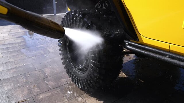 Bright yellow off-road vehicle with rugged tires. Water jet cleans muddy tread with force. Clean water sprays across pavement surface. Vehicle gleams under sunlight