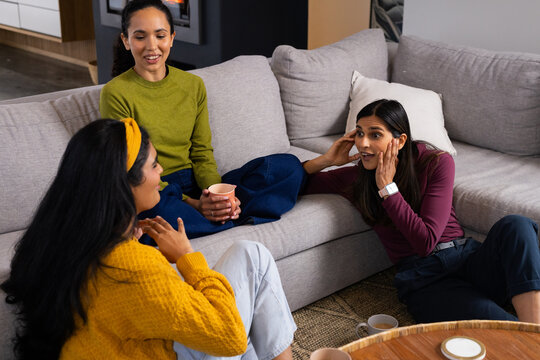 Diverse female friends sitting on sectional sofa and floor at home, wearing sweaters holding mugs
