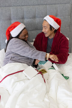 Couple Senior Asian man, African American woman sharing christmas gifts on bed wearing Santa hats