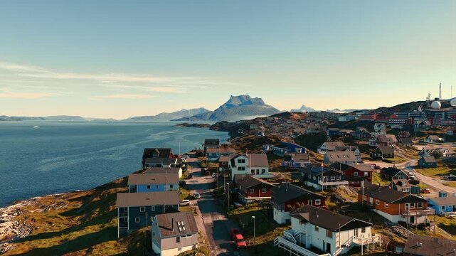 Cinematic drone footage of Nuuk, Greenland, captured during sunset, revealing colorful houses stretching along the Arctic coastline. Warm evening light reflects across the sea. 