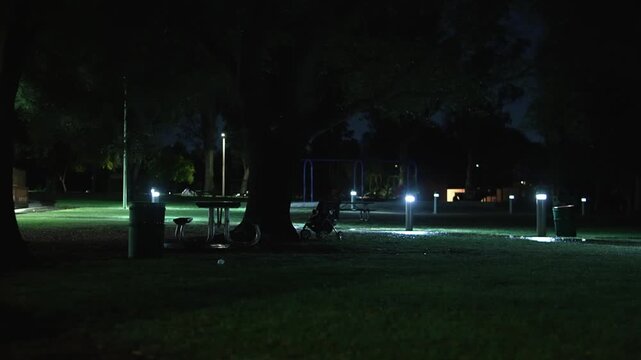 Empty playground and abandoned stroller in dark park at night are illuminated by cold pathway lights beneath large trees, creating a mysterious atmosphere