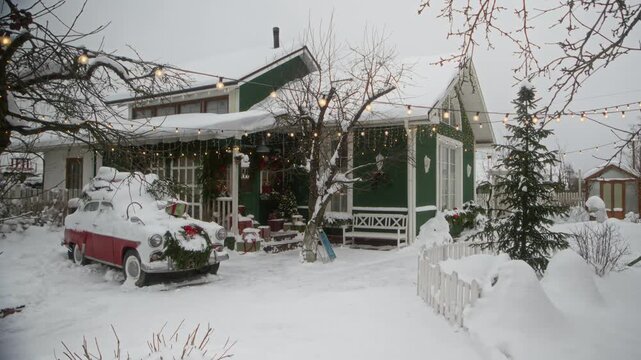 Illuminated string lights glowing on snow covered house facade with vintage car in winter yard. Establishing wide shot suitable for Christmas holiday advertising and seasonal travel campaigns