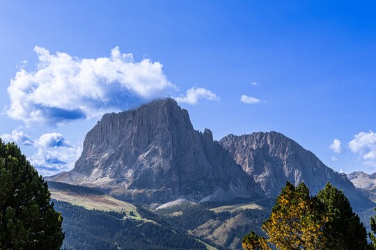 Massive rocky peak rises against vivid blue sky. Fluffy clouds drift gently near jagged summit. Green foliage frames foreground with autumnal tones. Mountain dominates view with quiet
