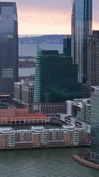 Vertical video: Purple sunset illuminating Exchange Place skyline, gleaming skyscrapers reflecting over Hudson River waters in Jersey City landscape