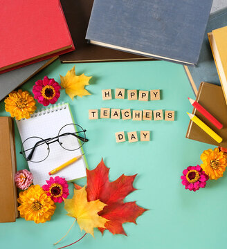 Teacher's day holiday, education concept. Text "Happy teacher's day" of wooden letters, notebook, pencils, learning books, eyeglass, flowers and autumn maple leaves on table. top view