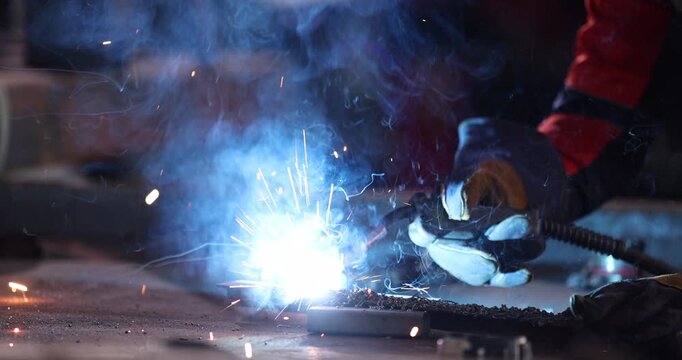 Welder wearing a protective welding helmet, gloves, and workwear performs a test weld on steel at a workbench.