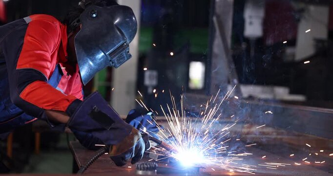 Welder wearing a protective welding helmet, gloves, and workwear performs a test weld on steel at a workbench.
