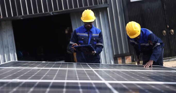 Two technicians wearing blue workwear and yellow hard hats inspect solar panels before installation, with one holding a digital tablet.