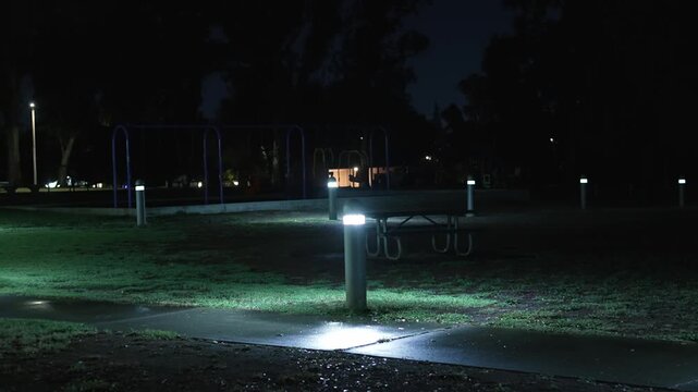 Empty playground at night illuminated by pathway lights after rain, with a wet walkway and deserted swings creating a mysterious and unsettling atmosphere