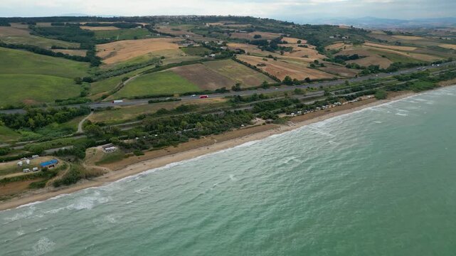 Aerial view of the Molise coast in Italy, showing a sandy beach along the turquoise Adriatic Sea. A coastal road, railway, highway, and seaside campsite border rolling agricultural fields and