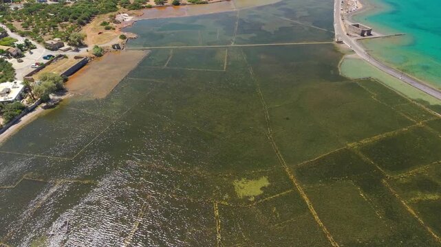 Submerged ancient ruins of Olous in Elounda, Crete, showing stone walls beneath shallow turquoise water next to a coastal road, aerial view