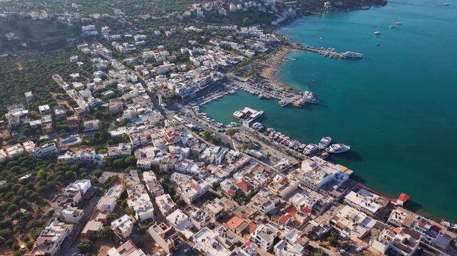 Aerial view of coastal town and harbor in Elounda, Crete, Greece, with boats moored along pier and turquoise Mediterranean water
