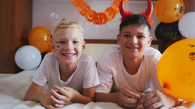 Two smiling boys with devil horns and halloween face paint look at camera lying on bed, slow motion. Concept of smiling kids enjoying halloween at home, great for party ads and social media