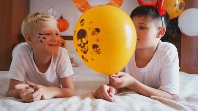 Boy with devil horns playing prank, holding skull balloon near friend face on bed, slow motion. Concept of playful halloween prank and kids mischief at home party, for ads, social media and blogs