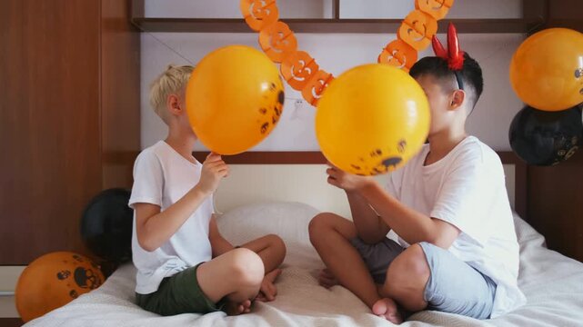 Two boys sitting cross legged on bed play with orange skull balloons, slow motion. Concept of active playtime and halloween party games for kids, playtime footage for ads, social media and blogs