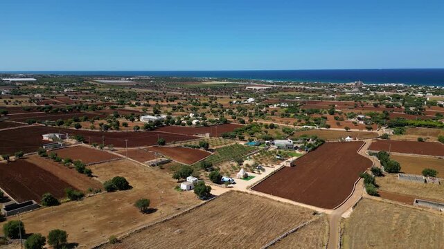 A high aerial view captures the rural Puglia countryside near Polignano a Mare, showcasing distinct red soil fields, scattered olive trees, and stone walls stretching towards the deep blue Adriatic