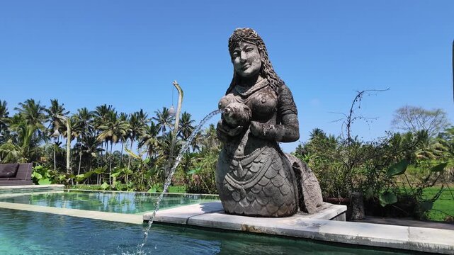 Stone statue fountain of a mythical figure in a tropical Ubud resort swimming pool, water splashing into pool, peaceful Balinese garden landscape, exotic travel, relaxation, luxury. Slow motion.