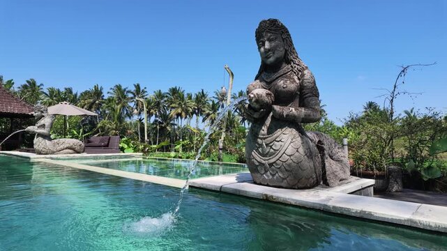 Stone statue of mermaid water fountain by swimming pool in Ubud Bali, tropical palm trees scenery, slow motion serene atmosphere for travel relaxation, spa wellness, resort luxury vacation, tranquil