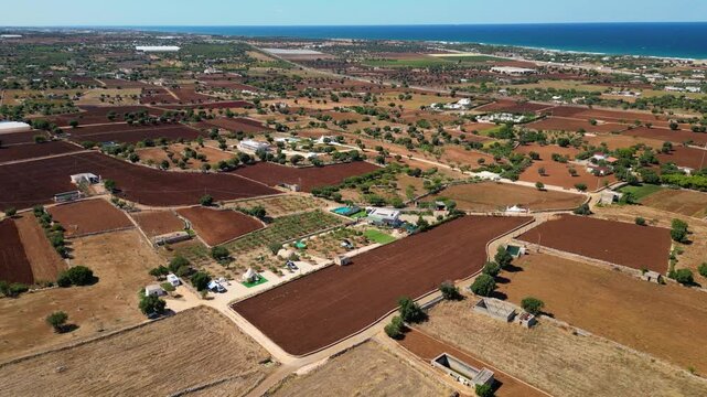 A high aerial view captures the rural Puglia countryside near Polignano a Mare, showcasing distinct red soil fields, scattered olive trees, and stone walls stretching towards the deep blue Adriatic