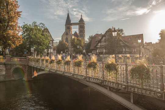 Panoramaansicht der St.-Agnes-Br&uuml;cke mit historistischen gusseisernem Gel&auml;nder, Stra&szlig;enlaternen  und Blumenschmuck im leuchtenden Seitenlicht vor der Stadtkirche St. Dionys in Esslingen am Neckar.