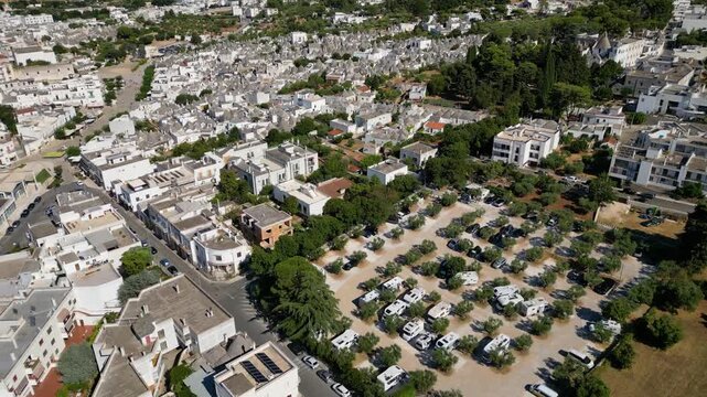 An aerial drone view shows a dedicated camperstop and RV parking area surrounded by olive trees in Alberobello, Italy. The facility sits conveniently right next to the historic UNESCO town filled with