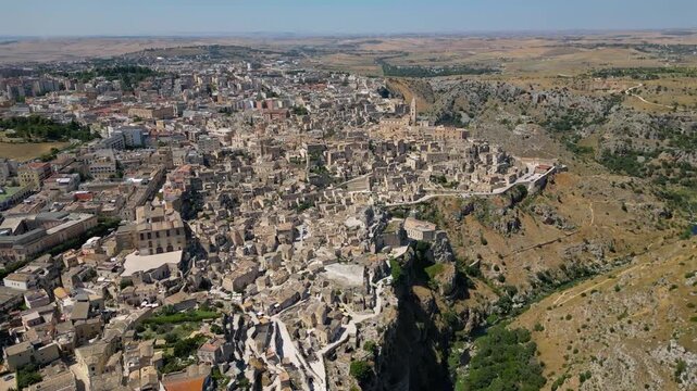 A high aerial panorama of Matera, Italy, showing the famous ancient Sassi quarter built along the edge of a dramatic canyon. The dense stone architecture, cathedral tower, and surrounding rugged