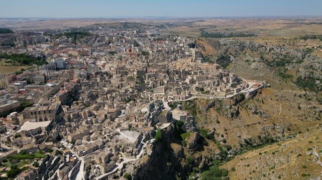 A high aerial panorama of Matera, Italy, showing the famous ancient Sassi quarter built along the edge of a dramatic canyon. The dense stone architecture, cathedral tower, and surrounding rugged