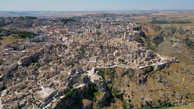 A high aerial panorama of Matera, Italy, showing the famous ancient Sassi quarter built along the edge of a dramatic canyon. The dense stone architecture, cathedral tower, and surrounding rugged