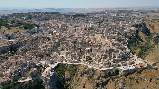 A high aerial panorama of Matera, Italy, showing the famous ancient Sassi quarter built along the edge of a dramatic canyon. The dense stone architecture, cathedral tower, and surrounding rugged