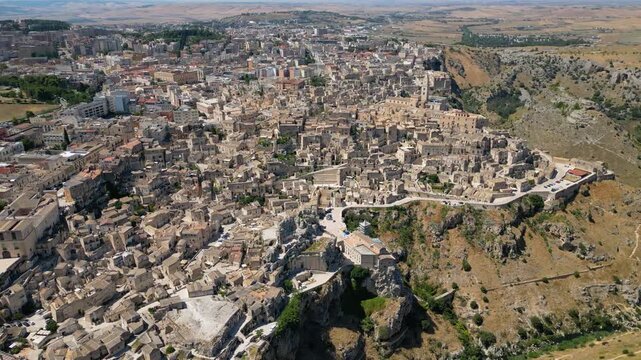 A high aerial panorama of Matera, Italy, showing the famous ancient Sassi quarter built along the edge of a dramatic canyon. The dense stone architecture, cathedral tower, and surrounding rugged