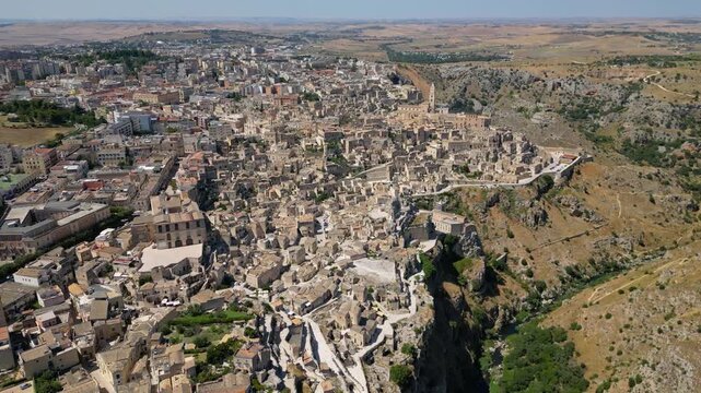 A high aerial panorama of Matera, Italy, showing the famous ancient Sassi quarter built along the edge of a dramatic canyon. The dense stone architecture, cathedral tower, and surrounding rugged