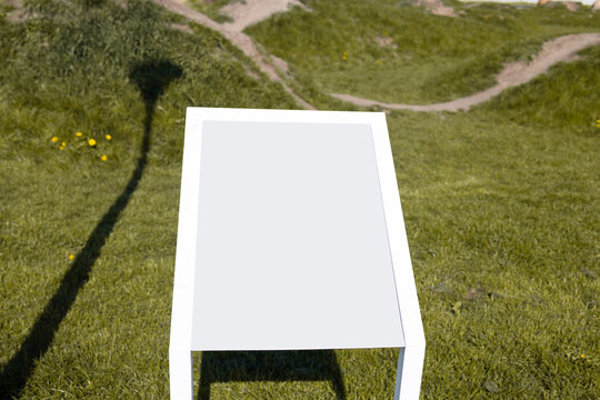 Contemporary white rectangular metal table or bench standing on a lush green lawn, with sharp shadows cast on the grass under bright sunlight. 