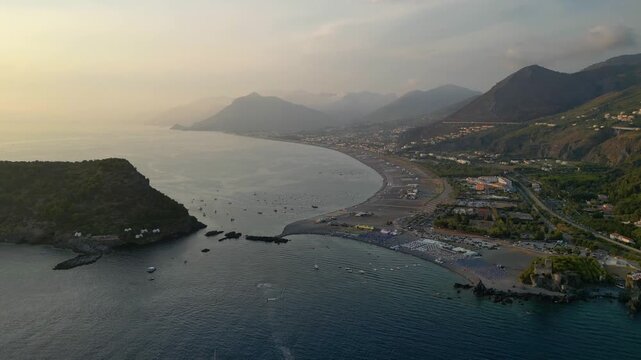 A breathtaking sunset aerial view captures Isola del Dino and the bay at Praia a Mare in Calabria, Italy. Boats navigate the deep blue waters along a sprawling beach, framed by dramatic coastal