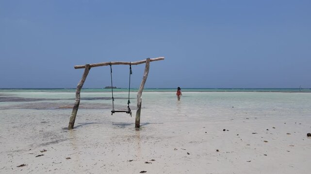 Wooden swing standing in crystal clear shallow tropical water with a woman walking in the distance, representing serenity, travel, summer vacation, island lifestyle, relaxing beach destination