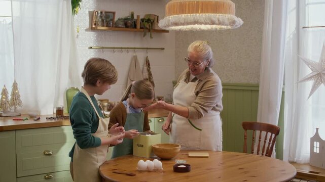 Senior Caucasian woman baking pastry with grandchildren at table while laughing playfully. Clip suitable for family lifestyle commercials, food brand campaigns and intergenerational cooking ads