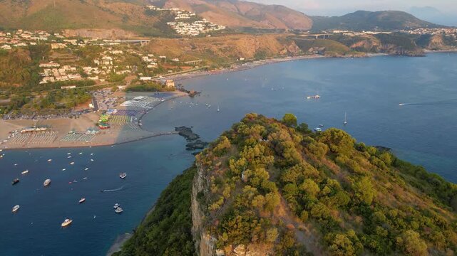 A breathtaking sunset aerial view captures Isola del Dino and the bay at Praia a Mare in Calabria, Italy. Boats navigate the deep blue waters along a sprawling beach, framed by dramatic coastal
