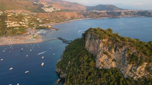 A breathtaking sunset aerial view captures Isola del Dino and the bay at Praia a Mare in Calabria, Italy. Boats navigate the deep blue waters along a sprawling beach, framed by dramatic coastal