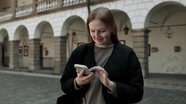Medium shot of a happy caucasian woman looking at her mobile phone screen and smiling, standing outdoors against a beautiful old town architecture with classic stone arches.