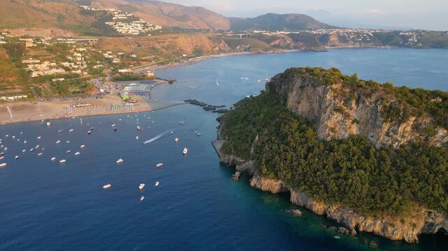 A breathtaking sunset aerial view captures Isola del Dino and the bay at Praia a Mare in Calabria, Italy. Boats navigate the deep blue waters along a sprawling beach, framed by dramatic coastal