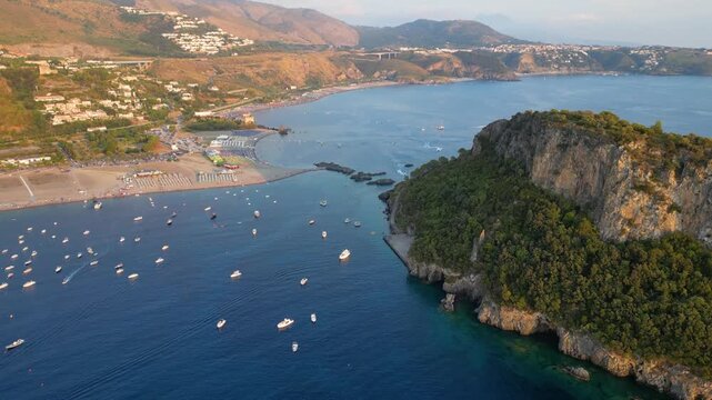 A breathtaking sunset aerial view captures Isola del Dino and the bay at Praia a Mare in Calabria, Italy. Boats navigate the deep blue waters along a sprawling beach, framed by dramatic coastal