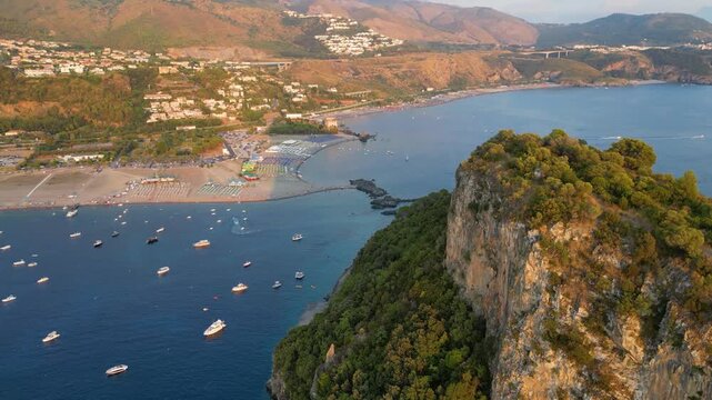 A breathtaking sunset aerial view captures Isola del Dino and the bay at Praia a Mare in Calabria, Italy. Boats navigate the deep blue waters along a sprawling beach, framed by dramatic coastal