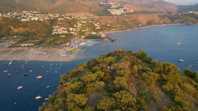 A breathtaking sunset aerial view captures Isola del Dino and the bay at Praia a Mare in Calabria, Italy. Boats navigate the deep blue waters along a sprawling beach, framed by dramatic coastal