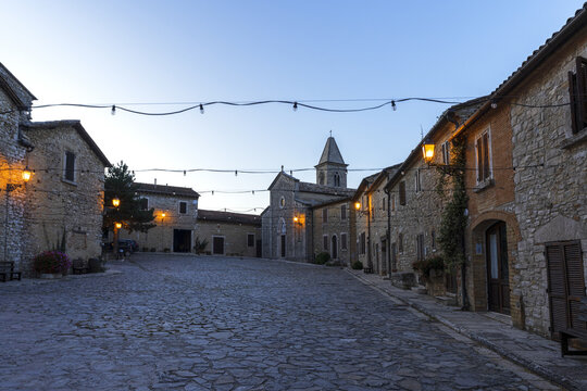 View of the historic square at Castello di Titignano with stone buildings, a church bell tower, and string lights at dusk in Orvieto, Umbria, Italy.