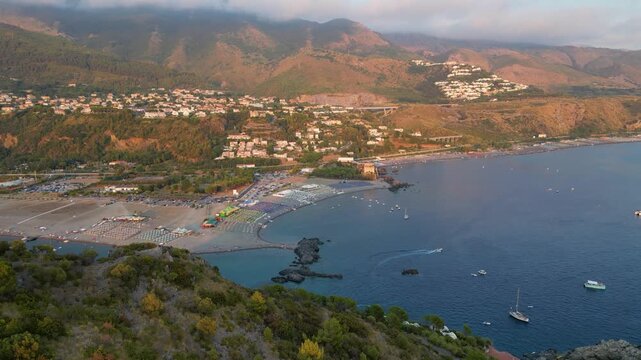 A breathtaking sunset aerial view captures Isola del Dino and the bay at Praia a Mare in Calabria, Italy. Boats navigate the deep blue waters along a sprawling beach, framed by dramatic coastal