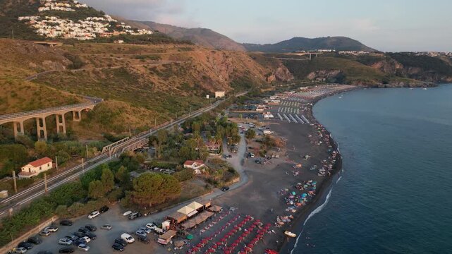 A breathtaking sunset aerial view captures Isola del Dino and the bay at Praia a Mare in Calabria, Italy. Boats navigate the deep blue waters along a sprawling beach, framed by dramatic coastal