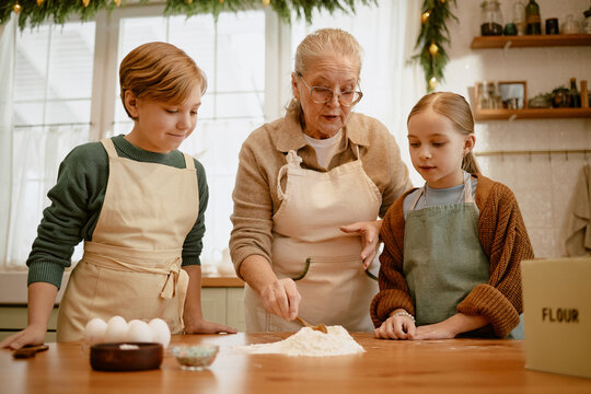 Senior Caucasian woman teaching Caucasian grandchildren baking in kitchen. Suitable for family lifestyle campaigns, holiday food marketing, and culinary workshops