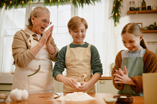 Senior Caucasian grandmother baking with grandson and granddaughter in kitchen, cracking egg into flour. Image ideal for family lifestyle campaigns, culinary advertising, food marketing