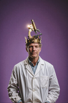 Man wearing a white lab coat stares forward while balancing a golden crown topped with an illuminated microscope on his head against a solid purple background.
